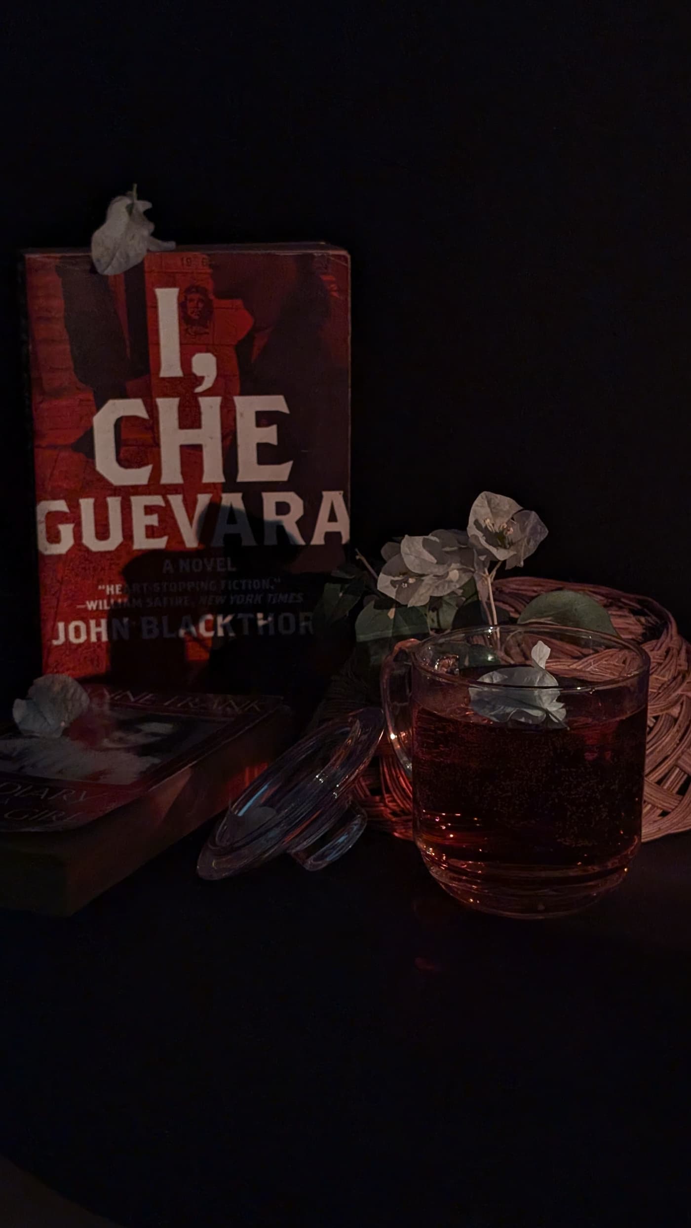 A hardback book, a glass of tea and dried flowers arranged on a dark surface, lit low and warm.