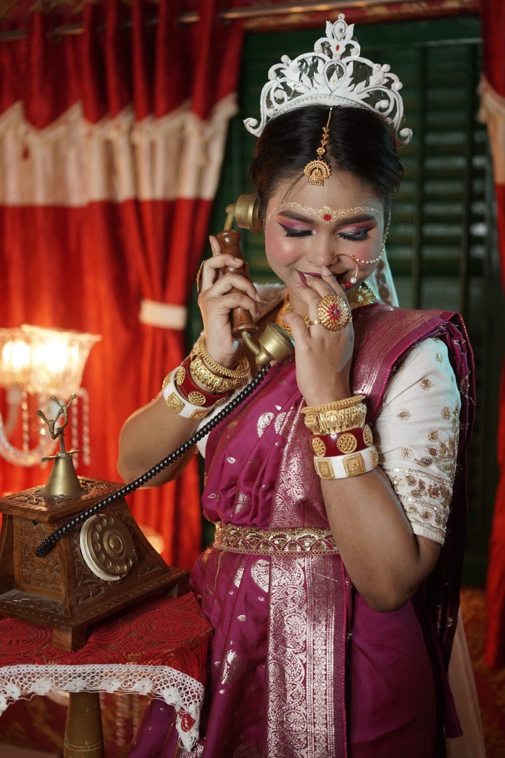 Sanjana Murmu in a magenta silk saree and silver mukut, smiling as she holds the receiver of a vintage brass telephone.