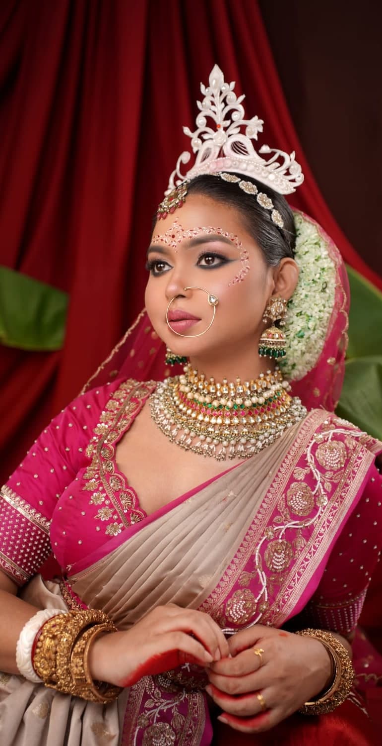 Sanjana Murmu in a magenta and gold bridal saree with a white crown and layered kundan necklaces, against a deep red curtain.